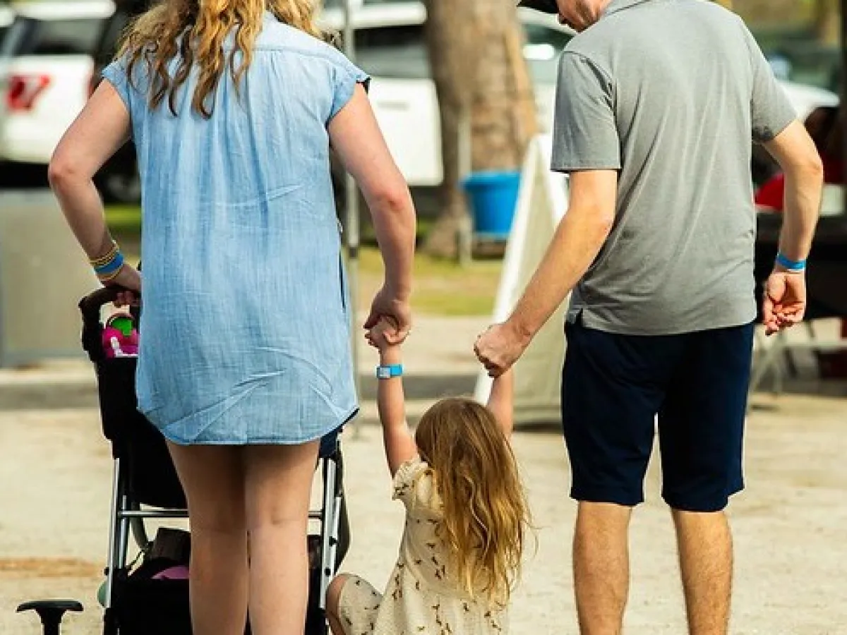 Two adults hold hands with a child swinging in between them, with a stroller nearby.